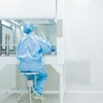 a scientist in full PPE sits in a cleanroom by an air hood working