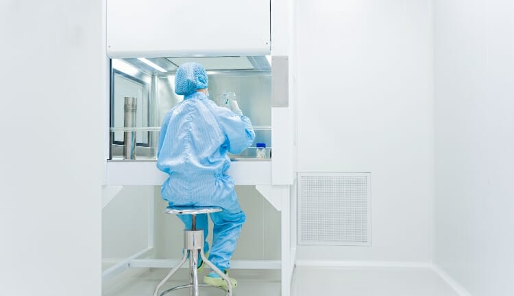 a scientist in full PPE sits in a cleanroom by an air hood working