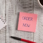 Handwriting text Order Now. Concept meaning the activity of asking for goods or services from a company Stationary placed next to a cup of black coffee above the wooden table.